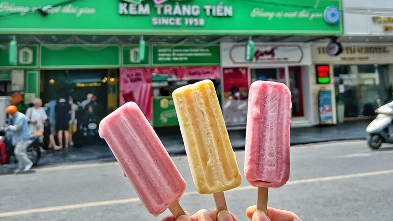 Một cây kem ốc quế Tràng Tiền vị vani đang chảy nhẹ, cầm trên tay, có biển hiệu Kem Tràng Tiền phía sau